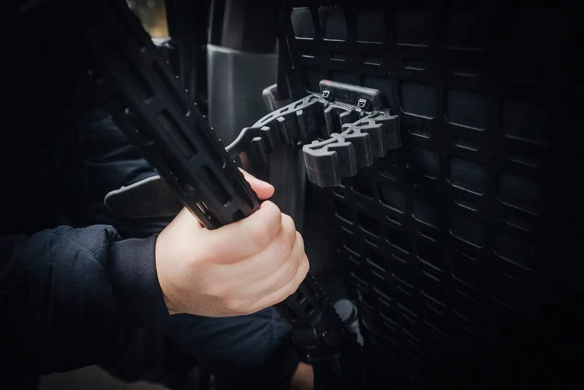 Руки фіксують гвинтівку AR вертикально на сталевій MOLLE панелі гумовими затискачами